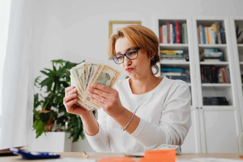 Woman wearing glasses, counting banknotes with a focused expression in a home office setting.